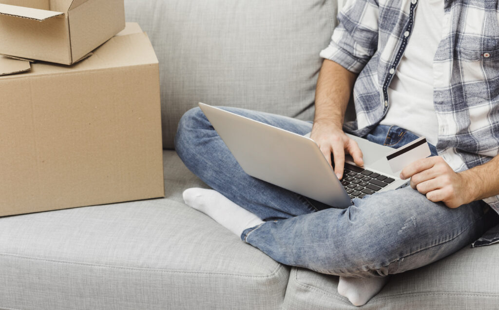 A man on a sofa holding a bank card and paying for an online order on his laptop, with parcels beside him.