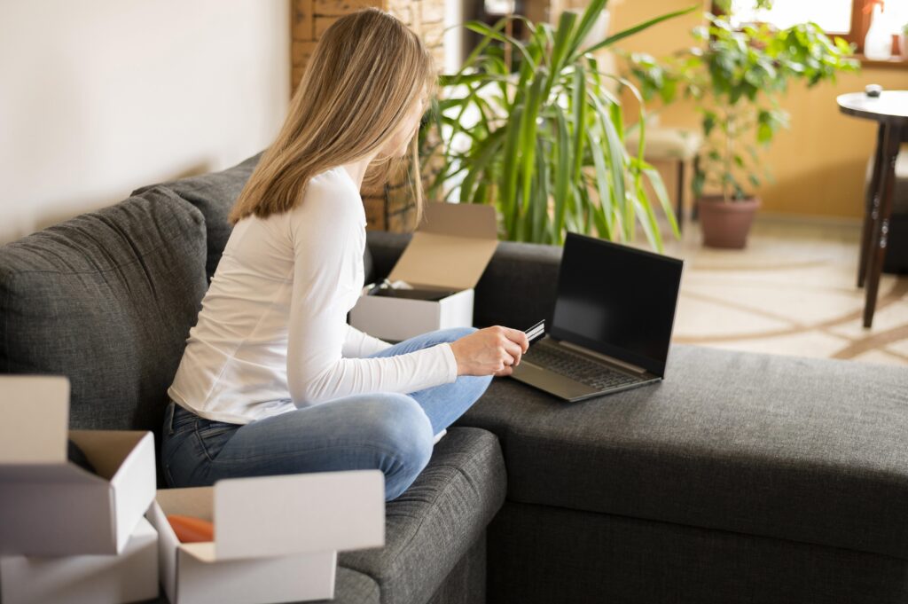 A woman shopping online on her laptop while sitting on a sofa, with open parcels beside her.